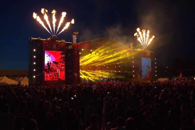 Eine große Menschenmenge auf einem Musikfestival schaut auf einer hell erleuchteten Bühne Performern zu, mit Zelten links und Feuerwerk, das den dunklen Himmel hinter beleuchtet.