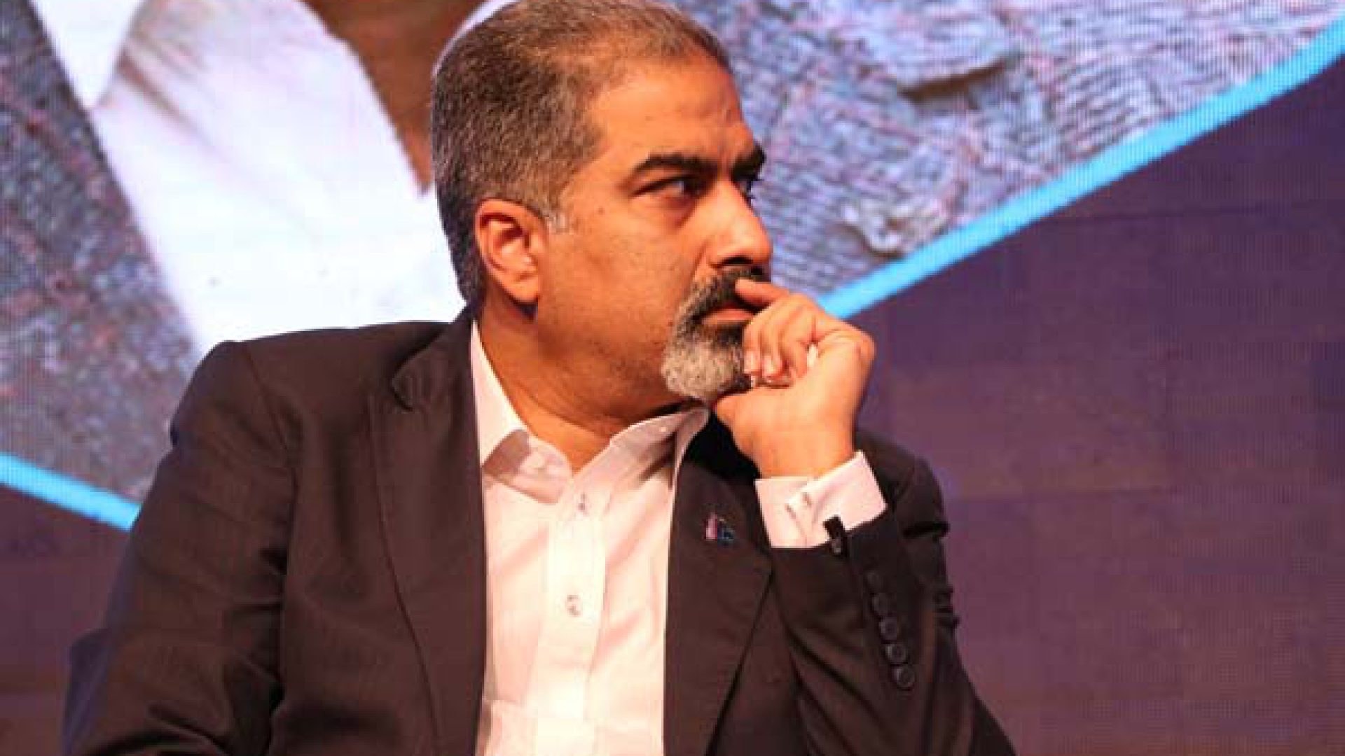 Ein Mann im Anzug und Krawatte sitzt auf einer Bühne und schaut nachdenklich auf einen Bildschirm.