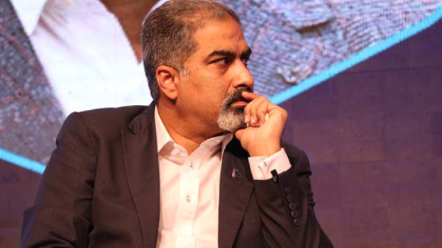 Ein Mann im Anzug und Krawatte sitzt auf einer Bühne und schaut nachdenklich auf einen Bildschirm.