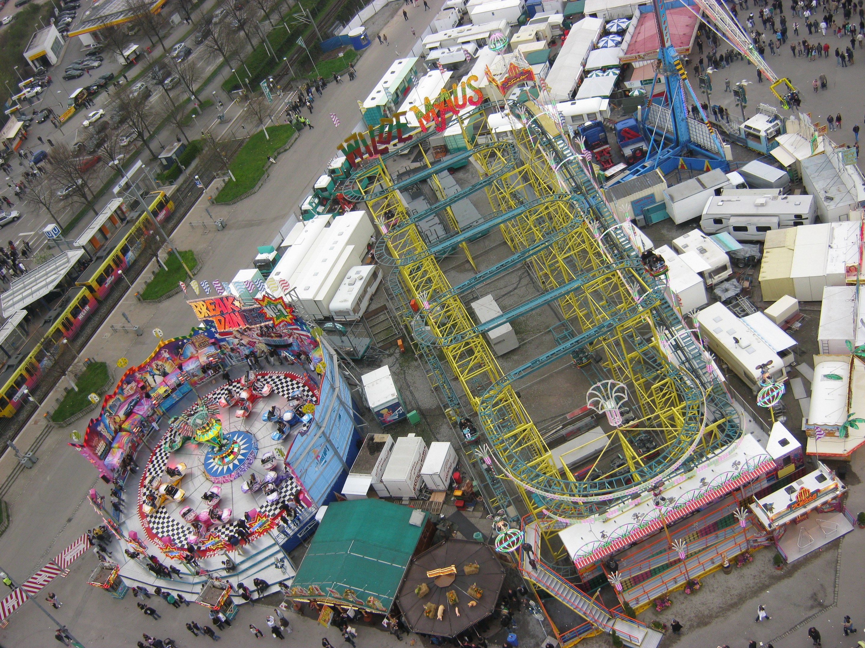 Luftaufnahme eines Vergnügungsparks mit einem zentralen Riesenrad, umgeben von verschiedenen Attraktionen, Gebäuden, Fahrzeugen, Menschen, Straßen und Vegetation.