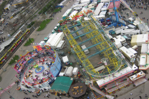 Luftaufnahme eines Vergnügungsparks mit einem zentralen Riesenrad, umgeben von verschiedenen Attraktionen, Gebäuden, Fahrzeugen, Menschen, Straßen und Vegetation.