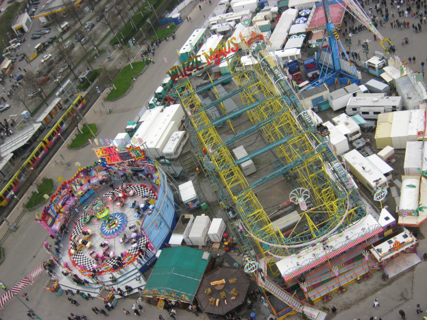 Luftaufnahme eines Vergnügungsparks mit einem zentralen Riesenrad, umgeben von verschiedenen Attraktionen, Gebäuden, Fahrzeugen, Menschen, Straßen und Vegetation.