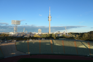 Olympiastadion in Berlin, Deutschland, mit dem Fernsehturm (Fernsehturm) im Hintergrund, umgeben von B"umen, Geb"uden und Lichtern unter einem bew"olkten Himmel.