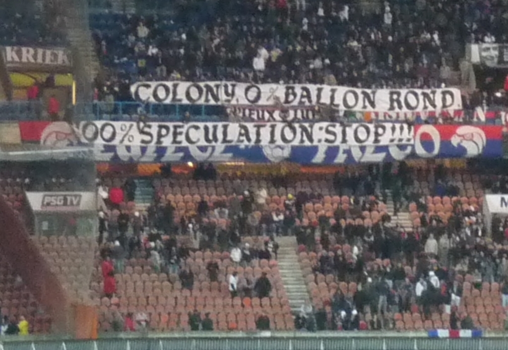 Ein Stadion voller großer Menschenmenge, Banner und Stühle, mit einem Zaun unten und Treppen auf der linken Seite.