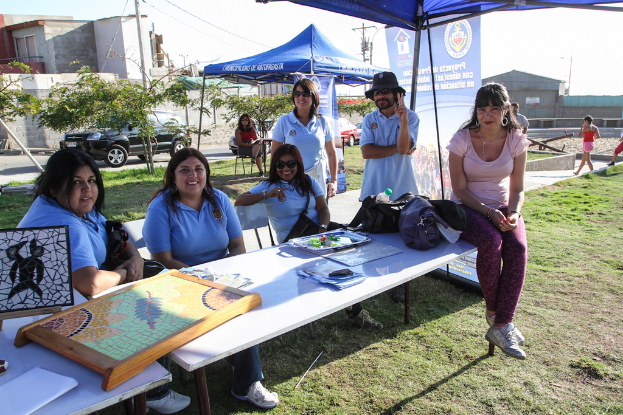 Eine Gruppe von Menschen sitzt um einen Tisch mit Spielen, Zelten und blauen Bannern im Hintergrund, während andere auf dem Gras stehen, ein Fahrzeug, Bäume, ein Haus und der Himmel sichtbar sind.