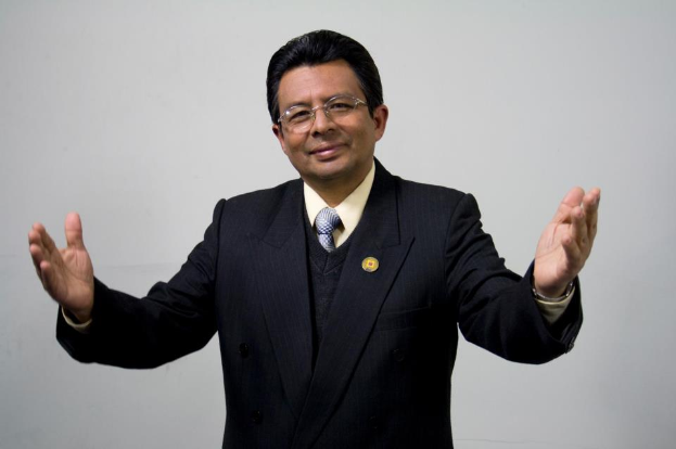 Ein Mann im Anzug und Krawatte mit ausgebreiteten Armen, der eine Brille trägt, steht vor einem weißen Hintergrund und sieht ernst aus.