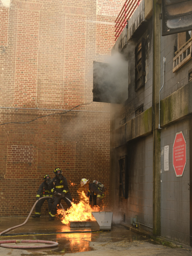 Feuerwehrleute in Helmen verwenden Schläuche, um ein Gebäude Feuer zu löschen, während Rauch aufsteigt; benachbarte Gebäude und ein Schild sind sichtbar.