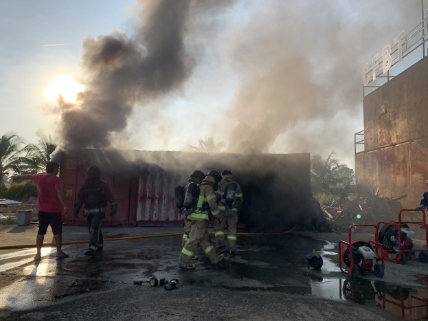 Eine Gruppe von Feuerwehrmännern in Helmen steht vor einem brennenden Gebäude mit aufsteigendem Rauch, während auf der rechten Seite ein Gebäude mit einer Namensplakette und links Bäume zu sehen sind.