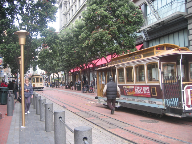 Eisenbahnschienen verlaufen entlang einer Straße mit einem Zug, der Text anzeigt, einer Person in der Nähe, kleinen Pfählen mit Seilen auf der linken Seite, Straßenlaternen, Bäumen, vielen Menschen im Hintergrund, einem Müllcontainer auf der linken Seite und Gebäuden mit Leitern auf der rechten Seite.