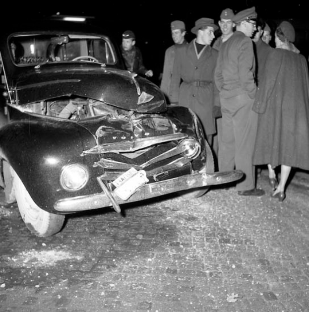 Ein Schwarz-Weiß-Bild eines beschädigten Autos am Straßenrand, umgeben von einer Gruppe von Menschen, die es besorgt betrachten.