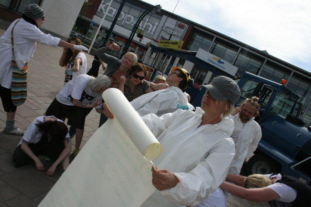 Eine Gruppe von Menschen, einige in Kostümen und Mützen, versammelt sich in der Mitte, wobei einige zu kämpfen scheinen, während eine Person ein Papier in den Vordergrund hält. Im Hintergrund sind Gebäude, Pflanzen, Pfosten und Schilder mit Text, ein Fahrzeug auf der rechten Seite, ein Gehweg unten und der Himmel oben zu sehen.
