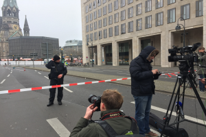 Eine Gruppe von Menschen steht auf der Straße in Berlin, ein Mann hält eine Kamera in den Vordergrund, Gebäude, Straßenlaternen, Autos, Absperrpoller, Schilder, Bäume und ein klarer blauer Himmel im Hintergrund.