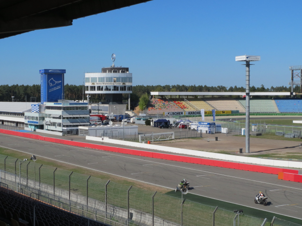 Eine Gruppe von Menschen auf Motorrädern auf einer Rennstrecke, umgeben von einem Metallzaun, Gras, Fahrzeugen, einem Gebäude, einem Schild, Pfählen, einem Stadion, Deckenleuchten, Bäumen und einem bewölkten Himmel.