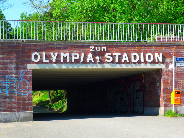 Der Eingang zum Olympiastadion in Berlin, Deutschland, mit einer Brücke mit Text, einem Metallzaun, einer Tafel, einem Karton, Pflanzen, Gras, einer Baumgruppe und einem bewölkten Himmel.