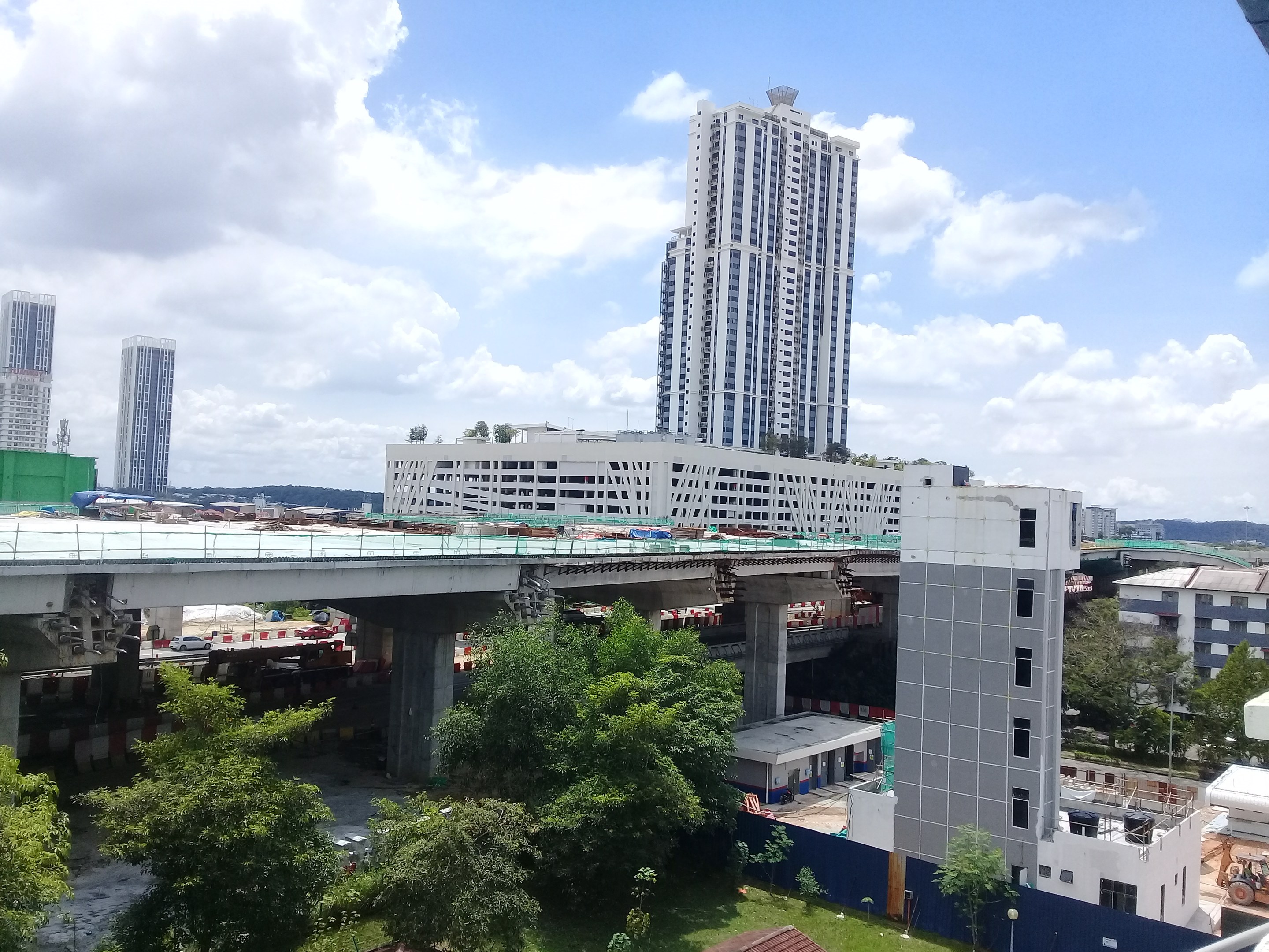 Luftaufnahme einer Stadt mit Gebäuden, Bäumen, Gras, Fahrzeugen, einer Brücke, Masten und bewölktem Himmel, die eine Baustelle für einen neuen Wohnkomplex zeigt.