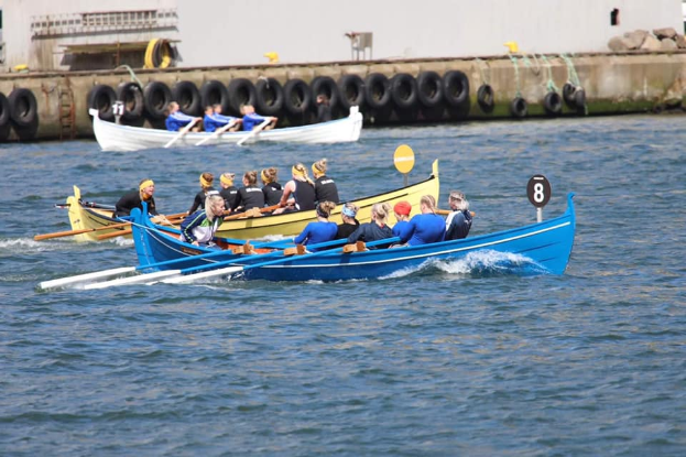 Eine Gruppe von Menschen in einem blauen und gelben Boot auf dem Wasser, die Paddel halten, mit einer Wand aus Reifen und einem Gebäude im Hintergrund, das auf eine Regatta hindeutet.
