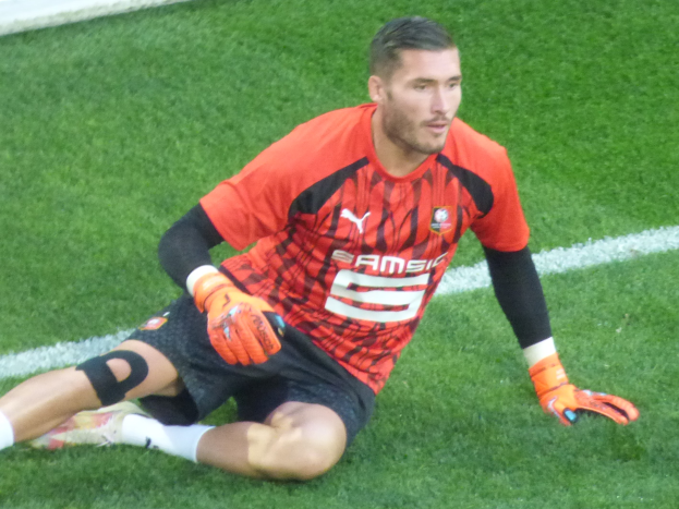 Ein Fußballspieler in rotem T-Shirt, schwarzen Shorts, weißen Socken und orangefarbenen Baseballhandschuhen sitzt auf dem Boden mit den Händen auf den Knien.