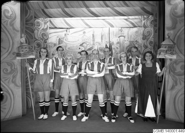 Eine Gruppe strahlender Frauen in 1940er-Kleidung steht auf einer Bühne und hält Stöcke in den Händen, im Hintergrund ist ein Gemälde zu sehen und unten steht der Text "1940er-Frauenfußballmannschaft".