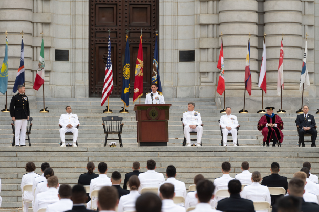 Gruppe von Menschen in weißen Uniformen auf Treppe eines Gebäudes sitzend mit Flaggen zu beiden Seiten eines Podiums vor einem großen Gebäude mit einer Tür.