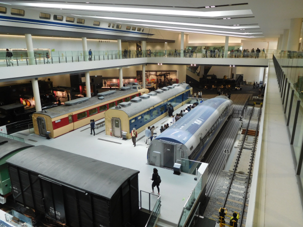 Ein geräumiger Museumsraum voller verschiedener Züge auf Display, Menschen beobachten, Geländer, Säulen und Deckenleuchten.