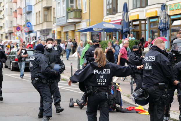 Eine Gruppe von Polizisten geht eine Straße entlang neben einer Menschenmenge, einige tragen Helme und Handschuhe, andere halten Schirme, mit Fahrzeugen, Schildern, Pflanzen und Gebäuden im Hintergrund.