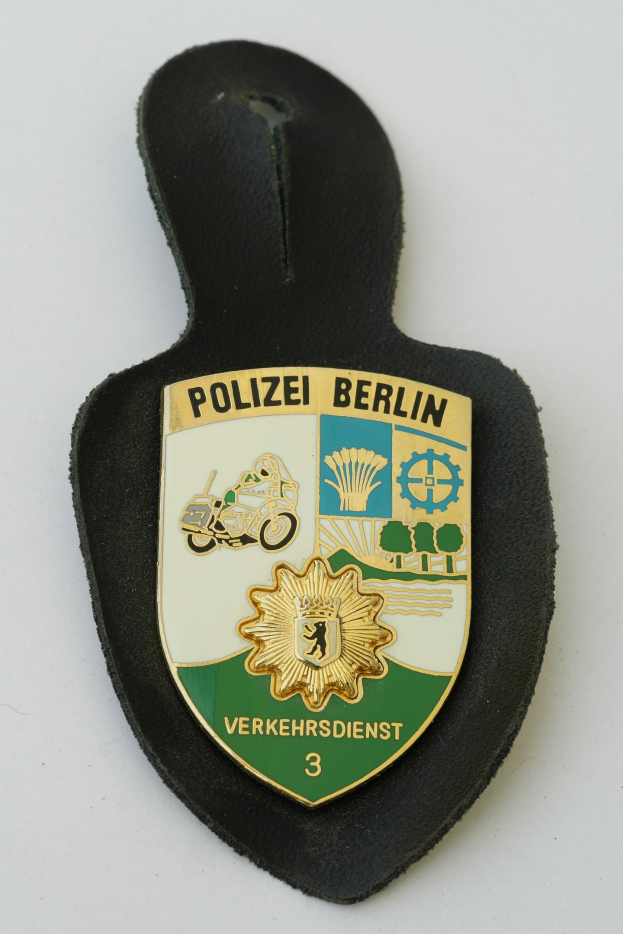 Ein schildförmiges Polizeibadged mit "Polizei Berlin" in schwarzer Schrift, das ein weißes Stern auf einem blauen Hintergrund und einen weißen Rand hat, liegt auf einer Oberfläche.