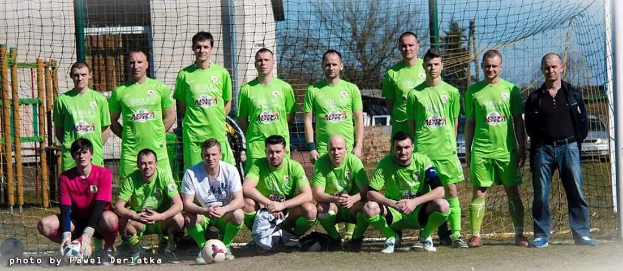 Gruppe von Männer in grünen T-Shirts auf einem Fussballfeld mit einem Ball und einem Tor, Bäume und Gebäude im Hintergrund.