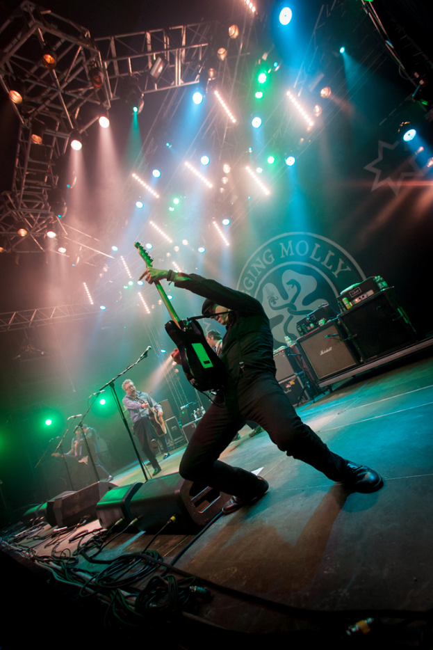 Ein musikalisches Konzert mit drei Künstlern auf der Bühne: zwei Gitarristen rechts mit Mikrofonen davor, ein Hintergrund mit 'Molly' darauf und zwei Schlangensymbole sowie Scheinwerfer darüber.