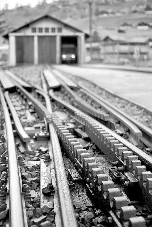 Eisenbahnschienen führen durch eine Szene mit einer Hütte, einem Fahrzeug und Häusern im Hintergrund.