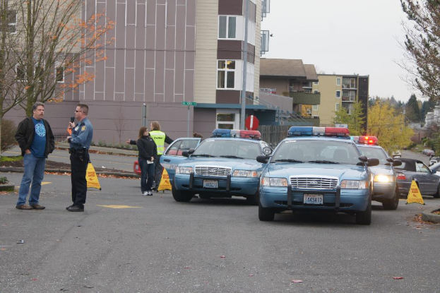 Autos auf einer Straße mit vier Menschen in der Nähe, Gebäude mit Fenstern im Hintergrund, Bäume und ein Notrufsignal.