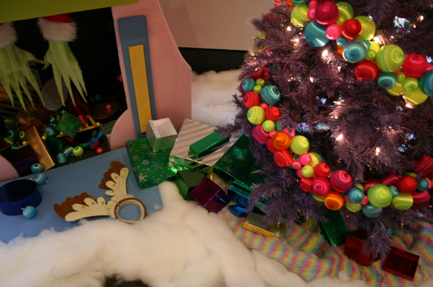 Drinnen steht rechts ein geschmückter Weihnachtsbaum mit Lichterketten und Schmuck, in der Mitte gibt es Geschenkboxen und Dekorationen, in der oberen linken Ecke eine Hütte mit Dekorationen und unten Wolle und Baumwolle.