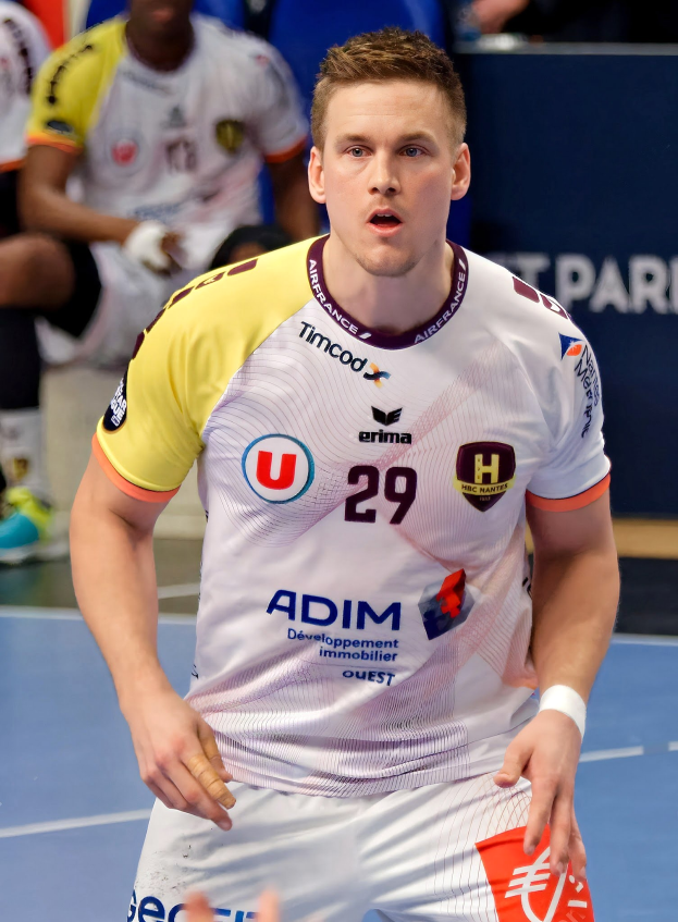 Ein Mann in einem weißen Hemd und gelben Shorts spielt Handball, hält ein Objekt und hat einen entschlossenen Gesichtsausdruck, während im Hintergrund Menschen auf dem Boden sitzen und eine Tafel die Aufschrift 'Ligue 1 - Futsal - Olympique de Marseille' trägt.