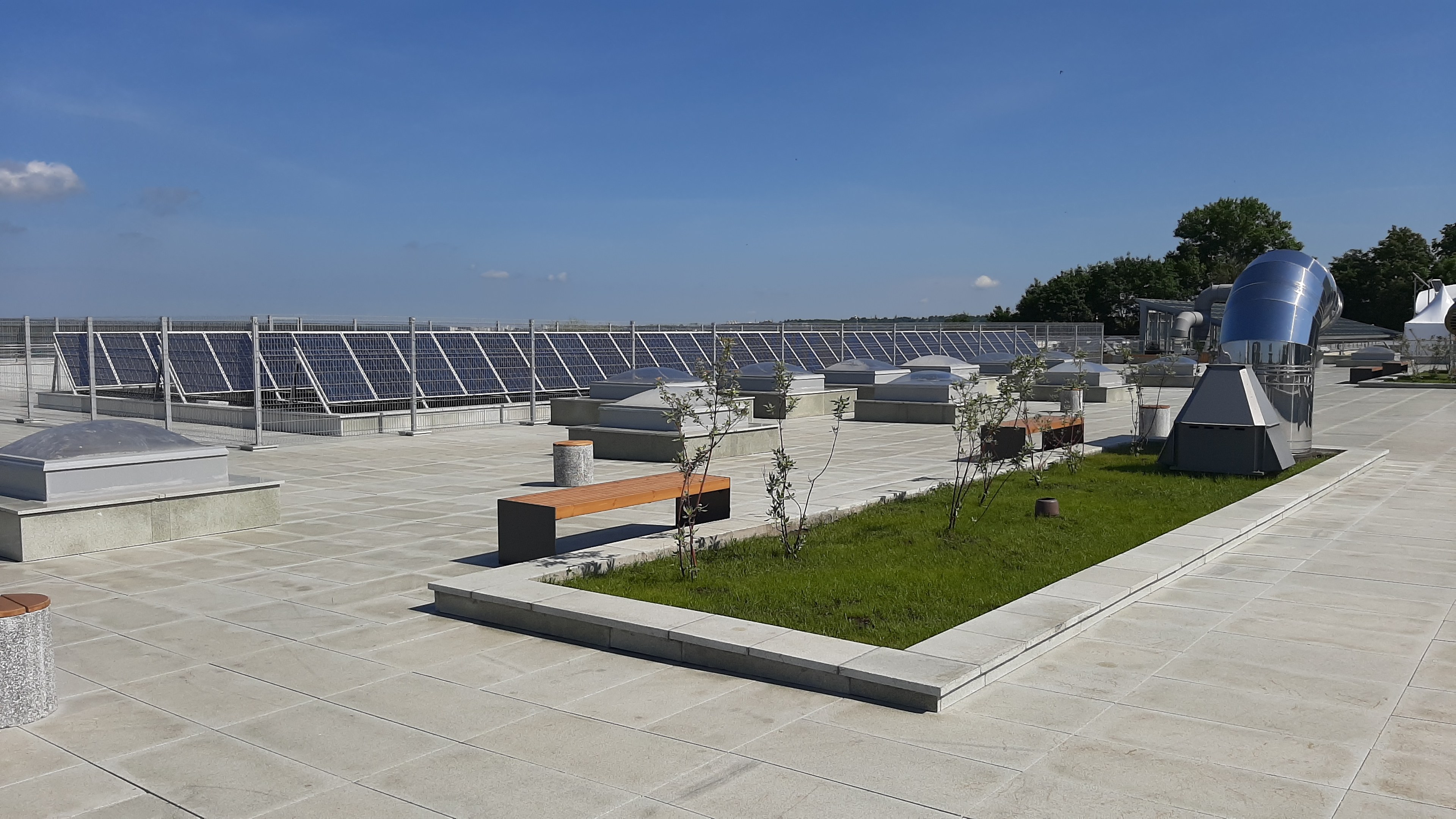 Gebäude mit Solarpaneelen auf dem Dach, umgeben von Bänken, Pflanzen, Gras und einem Hintergrund aus Bäumen und einer bewölkten Himmel.