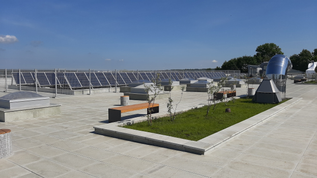 Gebäude mit Solarpaneelen auf dem Dach, umgeben von Bänken, Pflanzen, Gras und einem Hintergrund aus Bäumen und einer bewölkten Himmel.