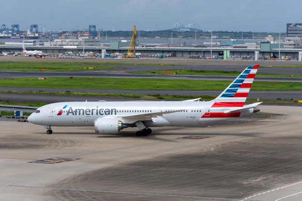 Ein American-Airlines-Flugzeug steht auf der Rollbahn eines Flughafens, umgeben von Gras, Fahrzeugen, Gebäuden, Masten, Kränen und anderen Gegenständen. Im Hintergrund gibt es Bäume und einen klaren blauen Himmel.