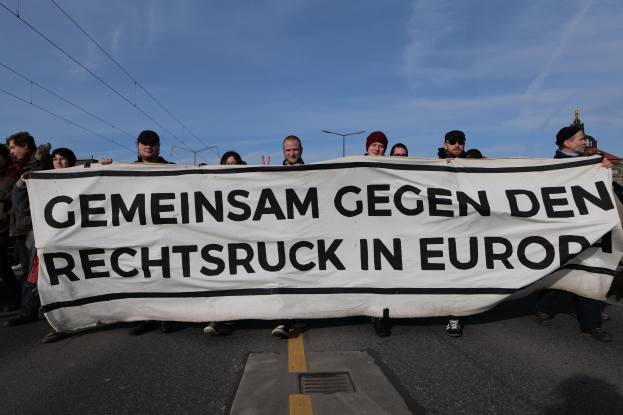 Eine Gruppe von Menschen auf der Straße mit einem Banner, auf dem "Gemeinsam Gegen den Rechtsruck in Europa" steht, vor Hintergrund von Straßenlaternen, Strommasten, Stromkabeln, Gebäuden und einem bewölkten Himmel.