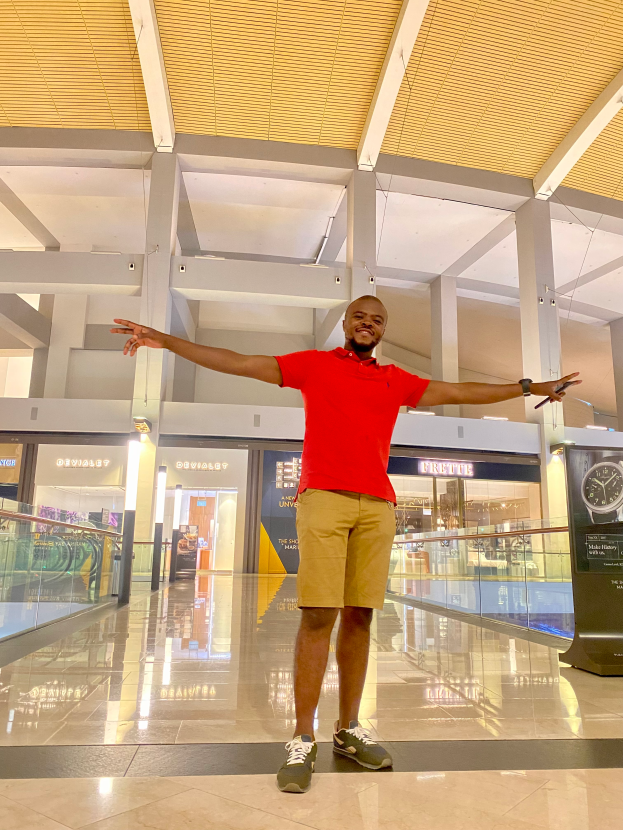 Ein Mann steht in einem Flughafen mit ausgebreiteten Armen, trägt ein rotes T-Shirt und cremefarbene Shorts, umgeben von Glaswänden, Säulen und Lichtern, mit einer Uhr auf der rechten Seite.