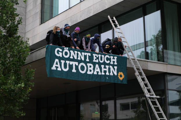 Gruppe von Menschen auf einem Gebäudedach mit einem Banner, einem Baum links und Glasfenstern im Hintergrund, die scheinbar für die deutsche Regierung protestieren.