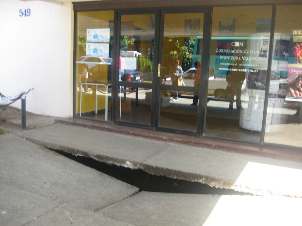 Eine gebrochene Oberfläche im Vordergrund mit einer Glastür, einer Wand und einem Pfosten im Hintergrund, die sich bewegende Fahrzeuge und einige stehende Personen spiegeln.