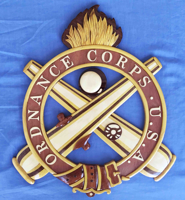 Holzschild mit "Ordnance Corps" in schwarzer Schrift, das ein weißes Stern auf einem navyblauen Hintergrund mit goldener Umrandung zeigt, auf blauem Tuch montiert.