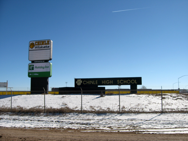 Eine Straße mit Schnee, ein Zaun mit Text, eine Wand, Laternenpfähle, Bäume und einen klaren Himmel, mit einem Schild im Vordergrund, auf dem "Chinle High School" steht.