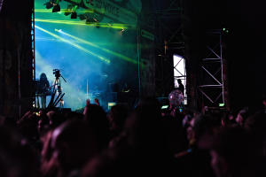 Eine Menge schaut Musikern auf einer hell erleuchteten Bühne mit Instrumenten, Mikrofonen und einem Banner bei einem Konzert in einem dunklen Veranstaltungsort zu.
