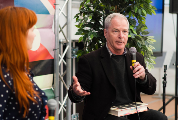 Ein Mann in einem schwarzen Blazer sitzt auf einem Stuhl und hält ein Mikrofon, mit einem Buch auf dem Schoß, während er einer Frau zu seiner Linken etwas erklärt, mit einer Fahne, einer Topfpflanze und einer Wand im Hintergrund.