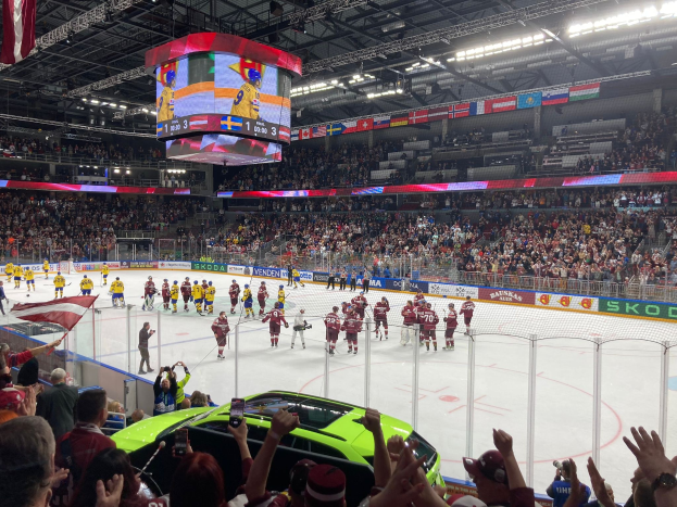 Ein Hockey-Spiel in einer großen Arena mit Zuschauern, ein Auto im Vordergrund, Banner, Deckenleuchten und hängende Fahnen.