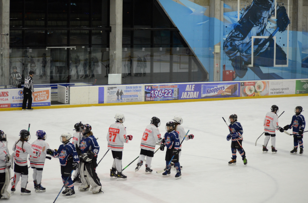 Gruppe junger Menschen mit Helmen und Hockeystöcken beim Eishockeyspielen auf einer Indoor-Eisfläche mit Bannern, Säulen und Glaswänden im Hintergrund.